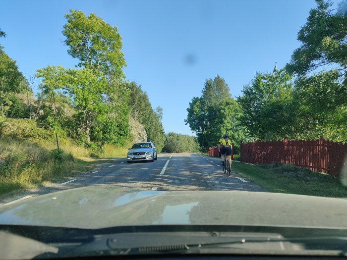Vy genom bilfönster visar en landsväg med en bil och en cyklist vid sidan under en klar himmel.