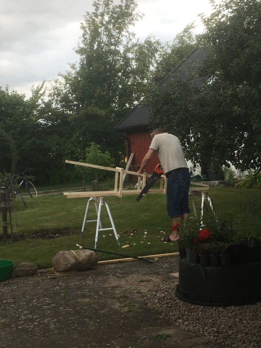 Person bygger växtstöd/prydnad i trädgård, ommålad stuga i bakgrunden.