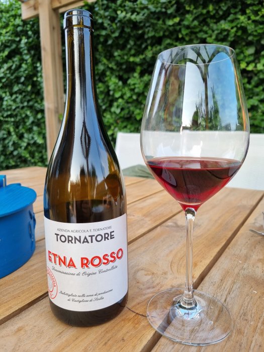 Flaska av Etna Rosso vin med logotyp bredvid ett glas med rödvin på ett träbord.