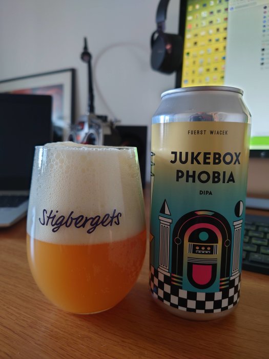 En ölburk märkt "Fuerst Wiacek JUKEBOX PHOBIA DIPA" bredvid ett ölglas med skummande öl på ett skrivbord.