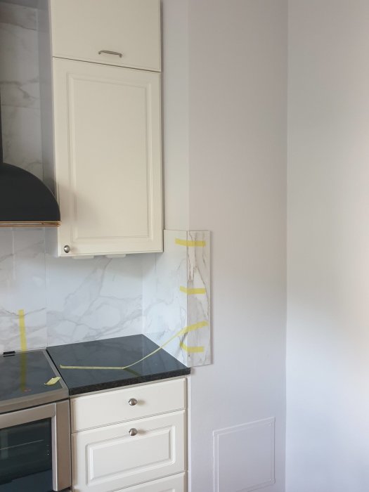 Kökshörn med marmorliknande tapet, vit skåpsinredning och en skiva som hörnskydd, tejpad för målning.