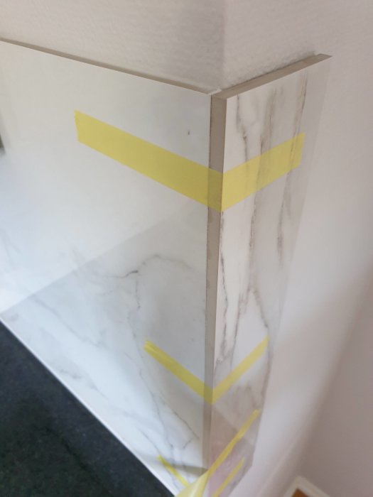 Hörn av vägg med marmormönstrade plattor och skyddande skiva, tejpade hörn som förberedelse för målning.