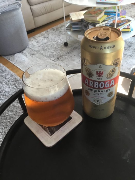 Ett glas öl och en ölburk av märket Arboga på ett svart bord, inomhusmiljö i bakgrunden.