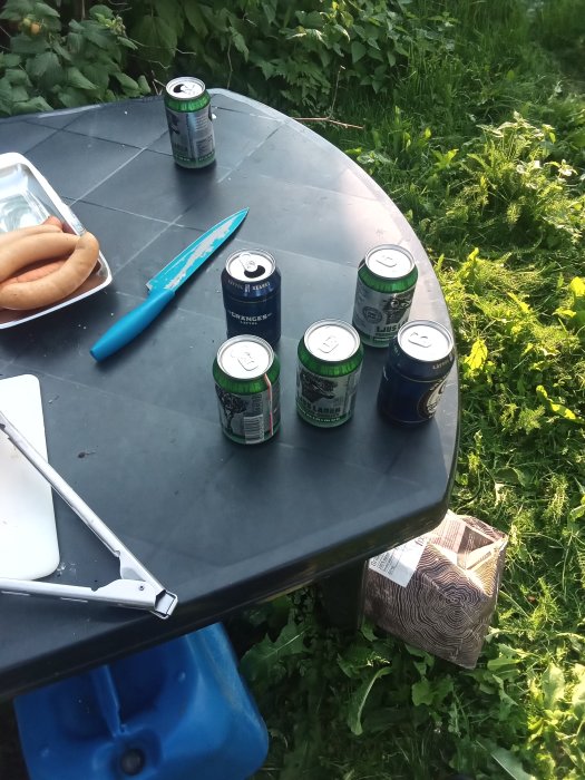 Lättölburkar på ett utebord med korv och grilltång, omgiven av grönska.