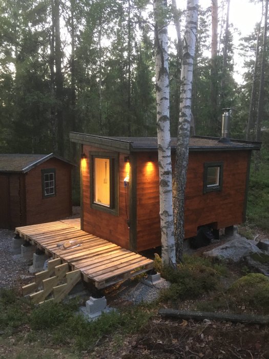 Bastustuga med tänd belysning och en veranda byggd av olika höga lastpallar i ett skogsområde vid skymningen.