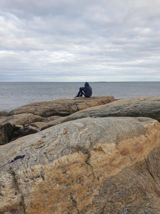 Person sittande på klippor vid vattnet med molnig himmel i bakgrunden.