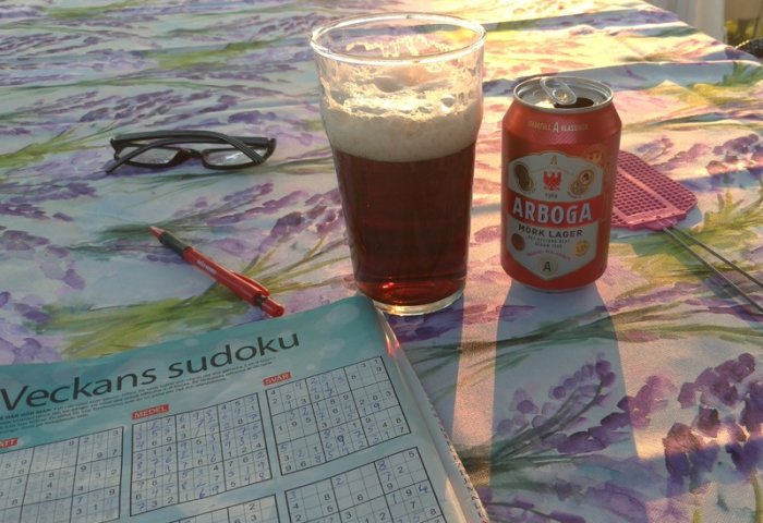 Sudoku-tidning, solglasögon, röd penna, ölglas med mörk lager och en burk Arboga öl på ett blommigt bord.