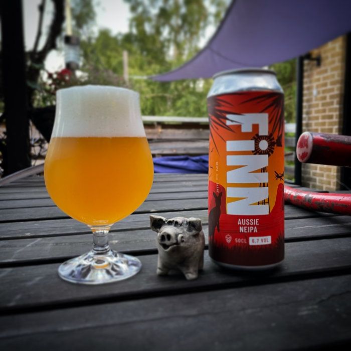 Ölglas fyllt med ljus öl, burk med etikett "Aussie NEIPA", och liten grisfigur på ett träbord utomhus.