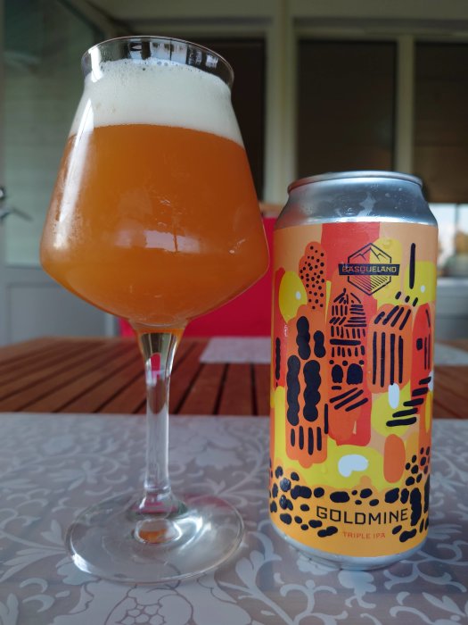 Ett glas fylligt öl bredvid en burk med etiketten "Basqueland Goldmine Triple IPA" på ett bord.