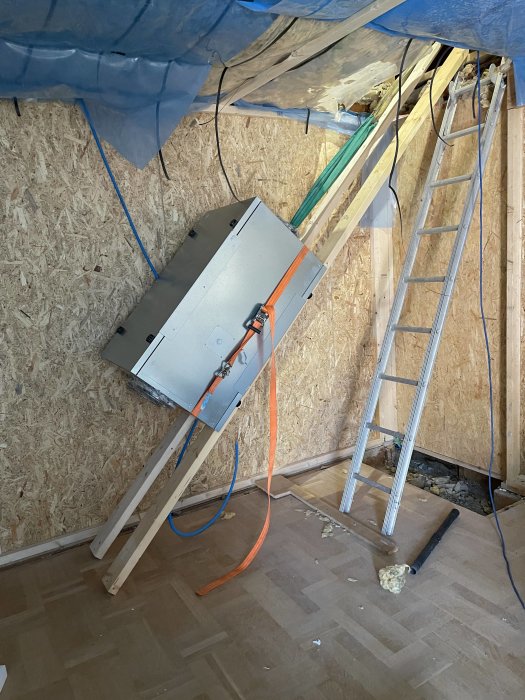 FTX-enhet upplyft på vinden med stöttande träslanor, nära en stege och oisolerat golv.