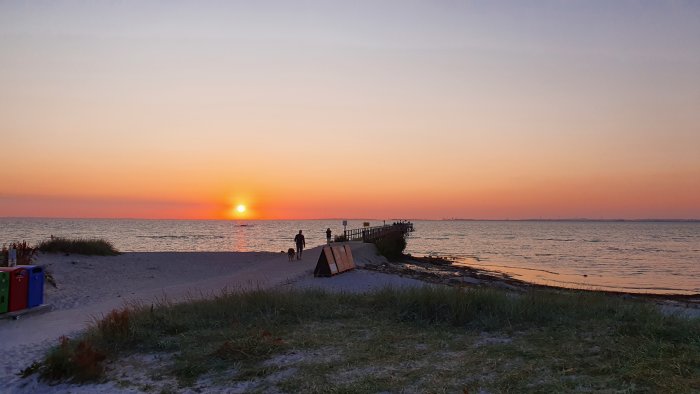 Solnedgång vid havet med människor på en pir och silhuett av stad i fjärran.