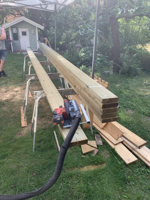 Brädor upplagda på bockar för sågning, med cirkelsåg, tvingar och dammutsugningsslang i trädgård.