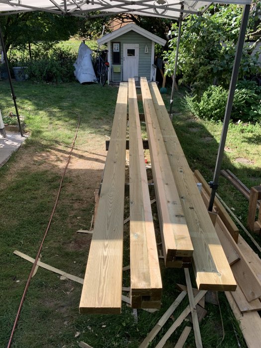 Långa träbrädor upplagda på bockar i trädgården med ett litet förrådshus i bakgrunden.