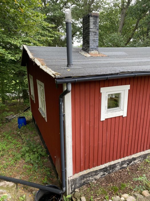 Rött trähus med svart tak som visar rötskador och behov av renovering, omgivet av grönska.