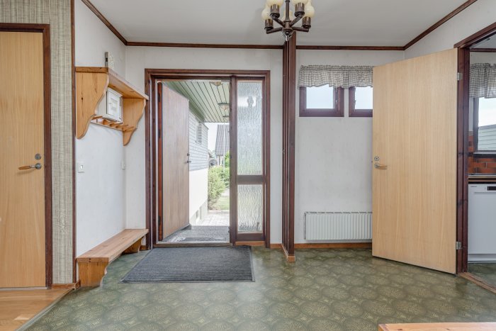 Ingångshall i hus från 1975 med öppen dörr, väggtelefon, trädetaljer och mönstrat golv.