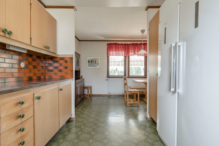 Odaterat kök med träskåp, tegelröd kakelvägg, mönstrat golv och bistrogardiner från 1970-talet.
