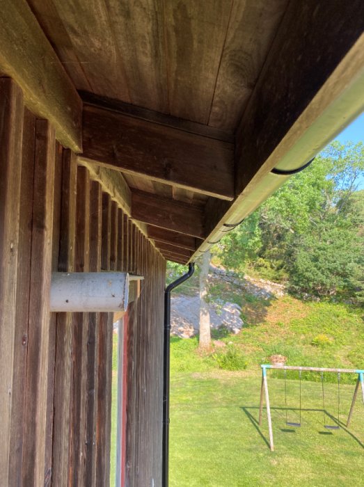 Avluftningsrör för avlopp på en yttervägg under husets takutsprång, med natur och en gungställning i bakgrunden.