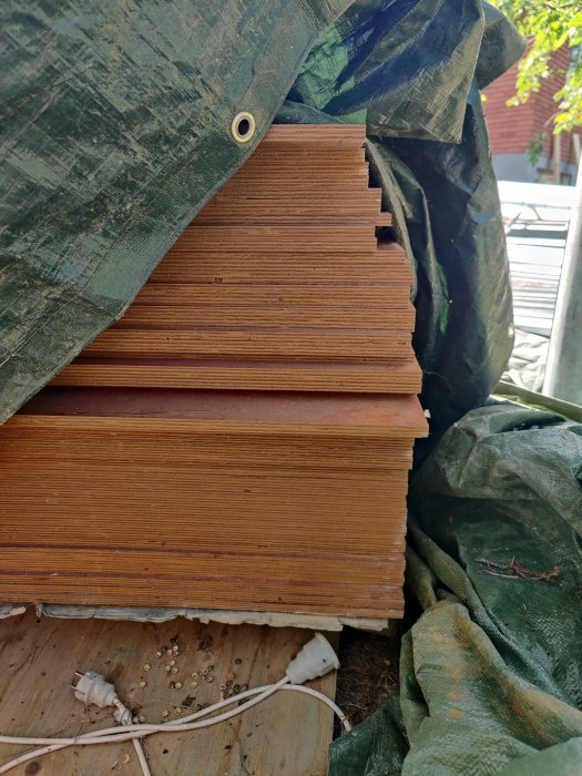 Stapel med formplywood skivor täckta med grön presenning på en träplattform.