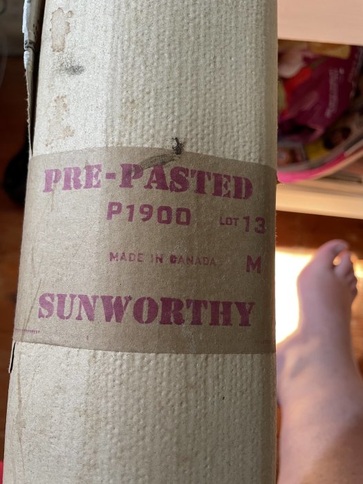 Förpackning av tapetrulle märkt "PRE-PASTED P1900" och "SUNWORTHY", gjord i Kanada, med en fot i bakgrunden.