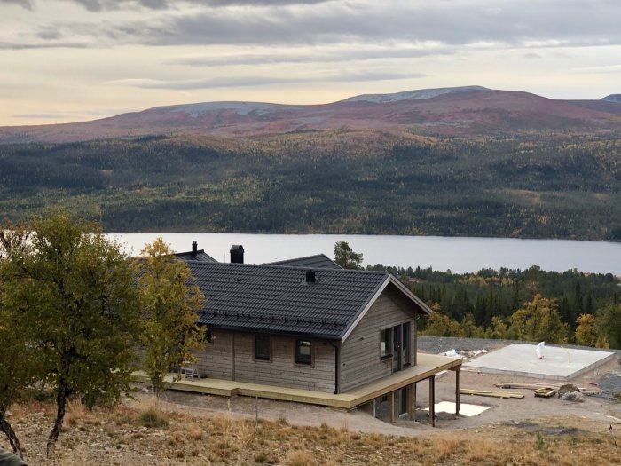 Fjällstuga med ljusgrå panel och svart tak framför ett bergslandskap och en sjö.