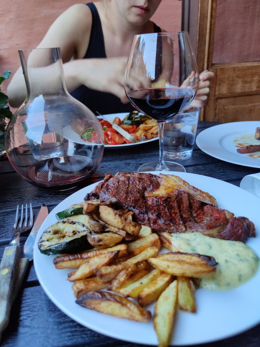 Middag med entrecote, klyftpotatis och grillade grönsaker på tallrik, rödvin i glas och karaff, person i bakgrunden.