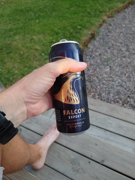 Hand håller en Falcon Export-ölburk utomhus på en trädäck efter en dag av snickeriarbete.