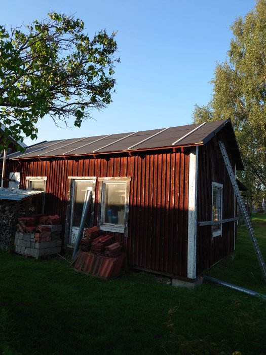 Reparerad hustomt med ny papp och ströläkt på taket, flankerad av byggmaterial och en stege.