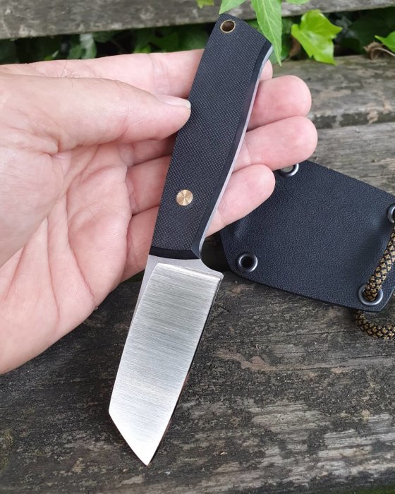 Hand som håller en handgjord kniv med svart handtag framför en träbänk och knivfodral.