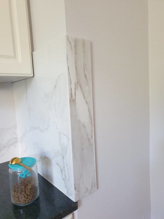 Marmormönstrad kakelplatta mot vit vägg intill köksskåp, med burk på bänkskiva.