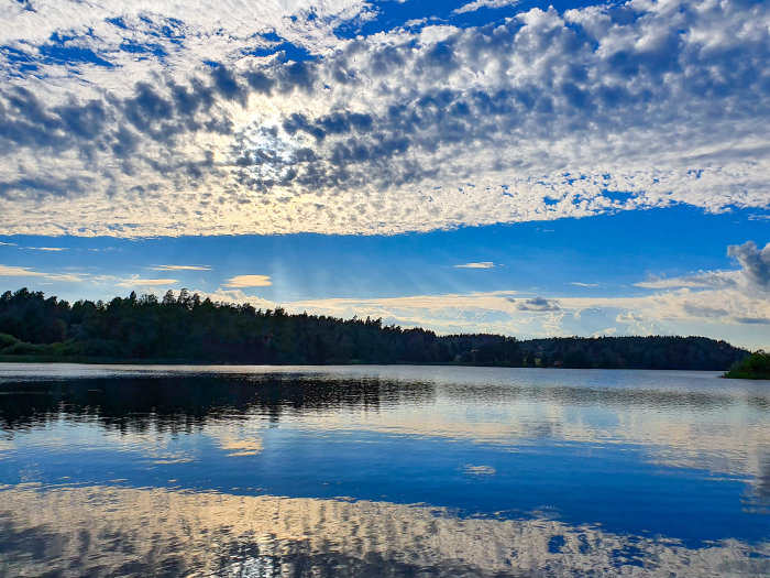 Kvällshimmel med moln reflekterade i en sjö omgiven av skog i början av juli.