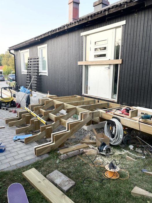 Pågående bygge av trätrappa utanför ett mörkfärgat hus med verktyg och byggmaterial utspridda på marken.