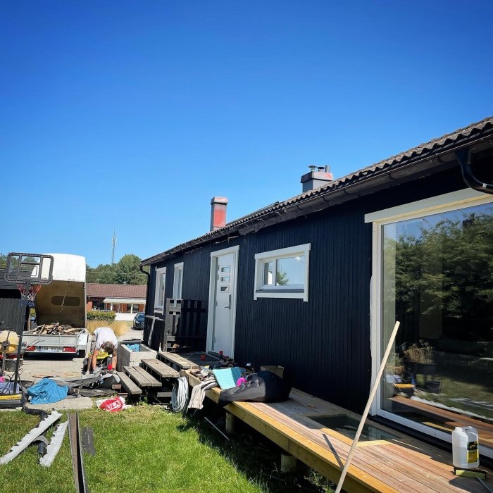 Byggarbetsplats med material och verktyg framför svart hus under en solig himmel.