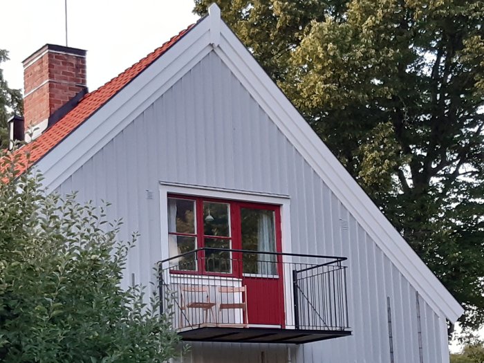 Nybyggd balkong på ett hus med vit fasad och röd dörr, omgiven av grönska.