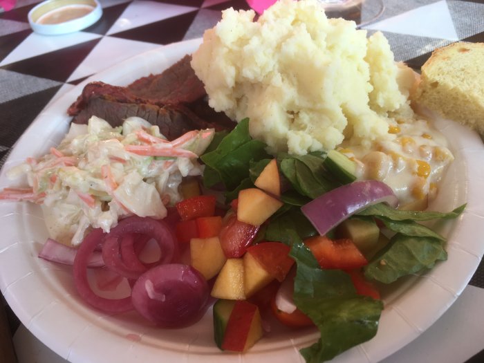 Tallrik med rökt brisket, potatismos, coleslaw, sallad, krämig majs, bröd och picklad rödlök.