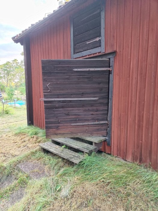 Röd träladugård med stora, gamla svarta dörrar och en borttagen sektion, omgiven av övervuxen gräs.