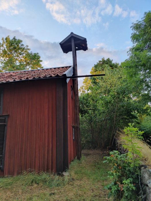 Gammalt rött trä hölager med svarta dörrar och en höftakstol, omringat av grönska och träd under en blå himmel.