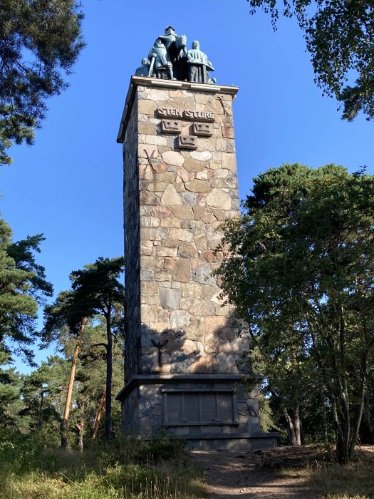Monument med inskriptionen "STEN STURE" omgiven av träd under blå himmel.