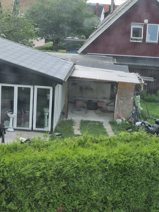 Utsikt över en trädgård med en utbyggnad mot ett rött hus, ej fristående, i ett villakvarter.