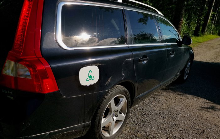 En svart Volvo V70 2,5FT från 2010 parkerad utomhus med en återvinningsdekal på tanklocket.