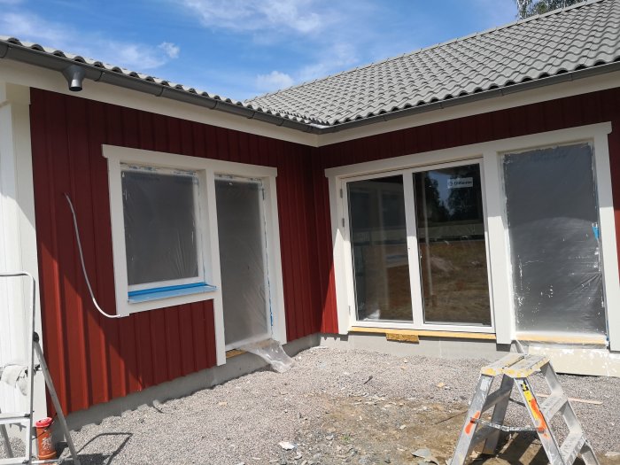Rödmålat hus med plasttäckt fönster och skyddad terrassdörr under målning, stege och målarburk syns.