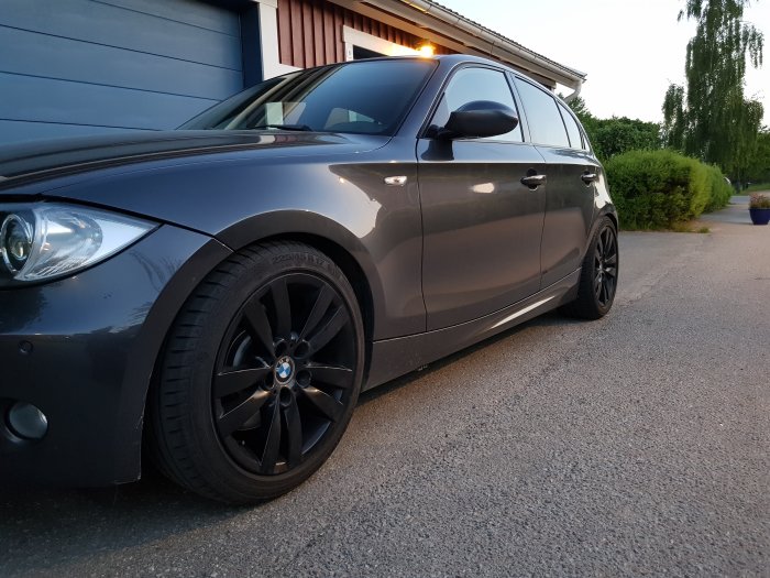 En svart BMW 130 parkerad vid en uppfart, kvällsbelysning, svarta fälgar.