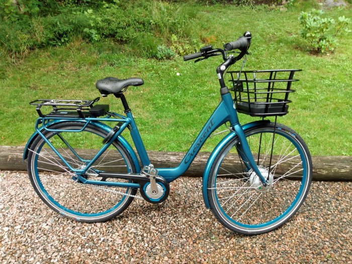 Turkos damcykel med korg parkerad på grus framför grönt buskage.