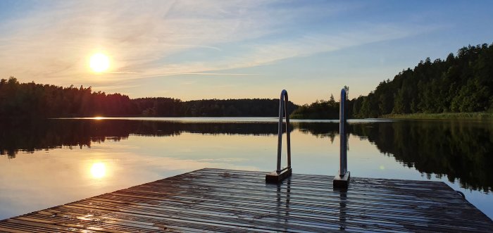 Solnedgång vid en sjö med en träbrygga och badstege, spegling av solen i vattnet.