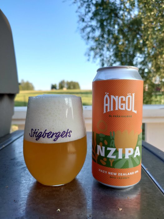 Ölglas med skum på bord utomhus bredvid en burk Ängöl NZIPA mot en bakgrund av landskap och träd.