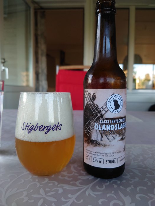 Ölflaska av Kackelbryggeriets Ölandslager bredvid ett glas med öl på ett bord med mönstrad duk.