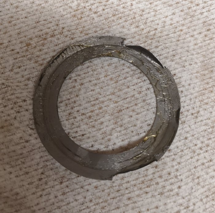Skadad metallisk shims med olja och synlig skada, placerad på en textil yta.
