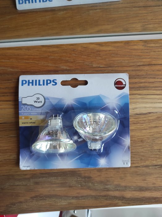 Förpackning av Philips 20 Watt, 205 lumen, dimbara halogenlampor med varmvitt ljus på ett träbord.
