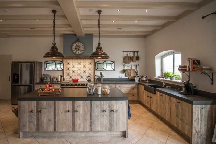Stilrent kök med rustika träskåp, svart bänkskiva, stora pendellampor och inbyggda vitvaror.