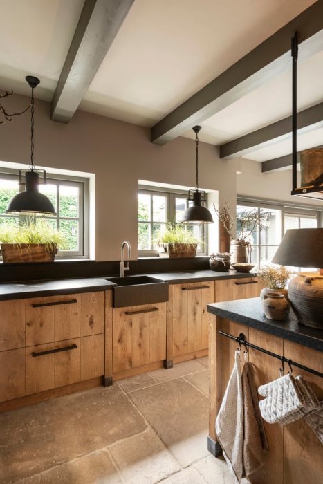 Stilfullt lantkök med träskåp, stenbänkskiva, industrilampor och fönster med gröna växter.