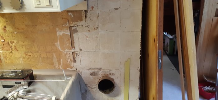 Ojämn köksvägg med spånskiva och murstock synlig, preparerad för renovering med synlig glipa nära diskbänken.